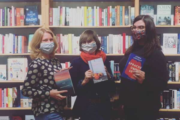 Das Team von Grimms Buchhandlung steht mit Buch in der Hand und Mundschutz vor einem Bücherregal.